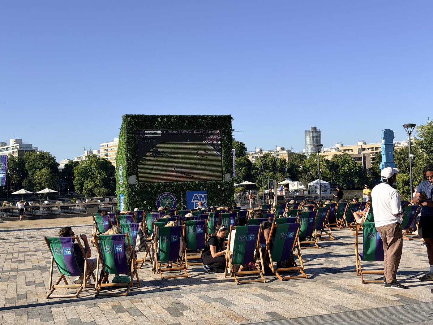 Deckchairs in a sunny square facing a big outdoor screen showing tennis